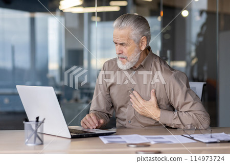 Older man, grey-haired, distressed, holding chest, experiencing pain while working on laptop in modern office setting with glass partitions and paperwork. Concept of health issues at work. Older man, grey-haired, distressed, holding chest, experiencing pain while working on laptop in modern office setting with glass partitions and paperwork. Concept of health issues at work. 114973724