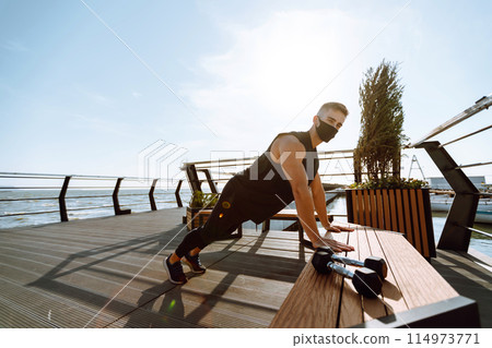 Handsome muscular man in protective mask, sports outfit doing functional exercises on seaside promenade. Handsome muscular man in protective mask, sports outfit doing functional exercises on seaside promenade. 114973771