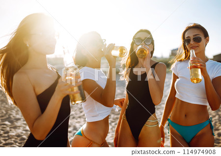 Four beautiful girls cheers and drink beers on the beach, enjoying vacation. Vacation time laughing and smiling. 114974908