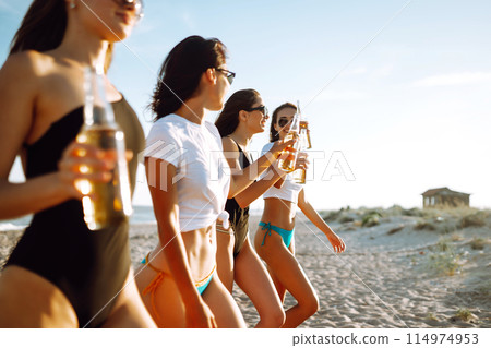 Four beautiful girls cheers and drink beers on the beach, enjoying vacation. Vacation time laughing and smiling. 114974953