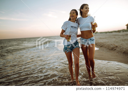 Beautiful two girls with glasses is holding a bottle with beer in her hands at the beach.. Summer lifestyle. Beautiful two girls with glasses is holding a bottle with beer in her hands at the beach.. Summer lifestyle. 114974971