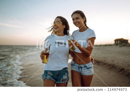 Beautiful two girls with glasses is holding a bottle with beer in her hands at the beach.. Summer lifestyle. Beautiful two girls with glasses is holding a bottle with beer in her hands at the beach.. Summer lifestyle. 114974973