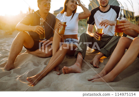 Young people sitting together at beach, drinking beer. Group of friends cheers with beers at the beach. 114975154