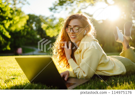 Young woman with laptop sits on grass in park on sunny day. Online training, remote work. Freelance. Young woman with laptop sits on grass in park on sunny day. Online training, remote work. Freelance. 114975176