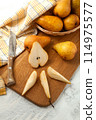 Cutting board with whole and slices pears, wicker basket and yellow kitchen towel on white wooden background.. 114975577