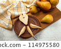 Clay plate with slices and half of pear, wicker basket and yellow kitchen towel on wooden cutting board.. 114975580