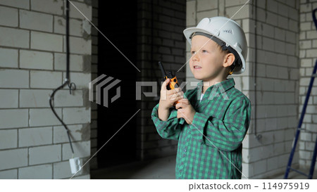 A small boy is standing on a construction site and talking on a walkie-talkie 114975919