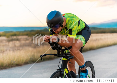 Close up photo of triathlete riding his bicycle during sunset, preparing for a marathon. The warm colors of the sky provide a beautiful backdrop for his determined and focused effort. 114977711