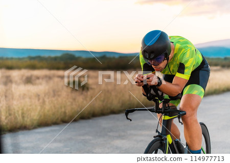 Triathlete riding his bicycle during sunset, preparing for a marathon. The warm colors of the sky provide a beautiful backdrop for his determined and focused effort. 114977713
