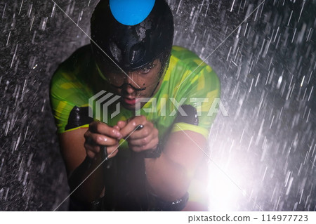 A triathlete braving the rain as he cycles through the night, preparing himself for the upcoming marathon. The blurred raindrops in the foreground and the dark, moody atmosphere in the background add 114977723