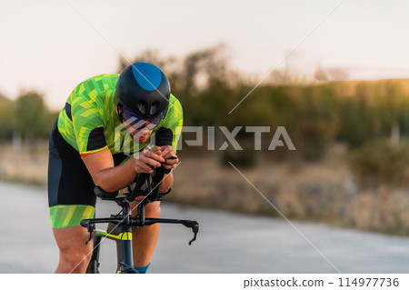 Triathlete riding his bicycle during sunset, preparing for a marathon. The warm colors of the sky provide a beautiful backdrop for his determined and focused effort. 114977736