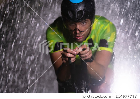 A triathlete braving the rain as he cycles through the night, preparing himself for the upcoming marathon. The blurred raindrops in the foreground and the dark, moody atmosphere in the background add 114977788