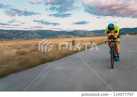 Triathlete riding his bicycle during sunset, preparing for a marathon. The warm colors of the sky provide a beautiful backdrop for his determined and focused effort. 114977797