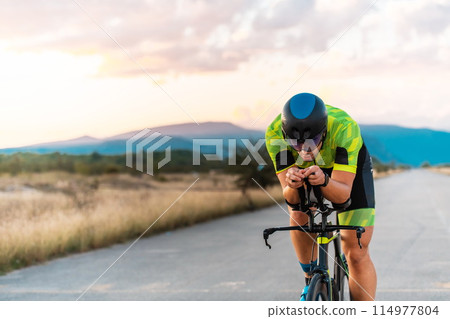 Triathlete riding his bicycle during sunset, preparing for a marathon. The warm colors of the sky provide a beautiful backdrop for his determined and focused effort. Triathlete riding his bicycle during sunset, preparing for a marathon. The warm colors of the sky provide a beautiful backdrop for his determined and focused effort. 114977804