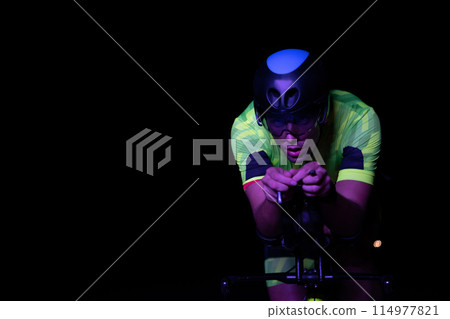 A triathlete rides his bike in the darkness of night, pushing himself to prepare for a marathon. The contrast between the darkness and the light of his bike creates a sense of drama and highlights the A triathlete rides his bike in the darkness of night, pushing himself to prepare for a marathon. The contrast between the darkness and the light of his bike creates a sense of drama and highlights the 114977821