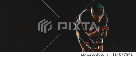 A triathlete rides his bike in the darkness of night, pushing himself to prepare for a marathon. The contrast between the darkness and the light of his bike creates a sense of drama and highlights the 114977841