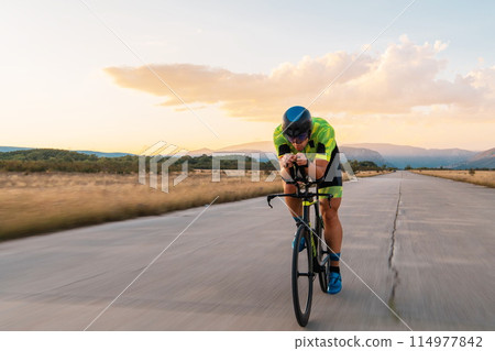 Triathlete riding his bicycle during sunset, preparing for a marathon. The warm colors of the sky provide a beautiful backdrop for his determined and focused effort. 114977842