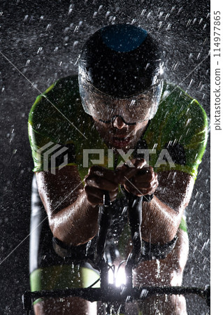 A triathlete braving the rain as he cycles through the night, preparing himself for the upcoming marathon. The blurred raindrops in the foreground and the dark, moody atmosphere in the background add A triathlete braving the rain as he cycles through the night, preparing himself for the upcoming marathon. The blurred raindrops in the foreground and the dark, moody atmosphere in the background add 114977865