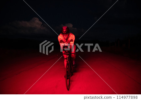 A triathlete rides his bike in the darkness of night, pushing himself to prepare for a marathon. The contrast between the darkness and the light of his bike creates a sense of drama and highlights the A triathlete rides his bike in the darkness of night, pushing himself to prepare for a marathon. The contrast between the darkness and the light of his bike creates a sense of drama and highlights the 114977898