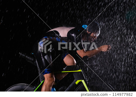 A triathlete braving the rain as he cycles through the night, preparing himself for the upcoming marathon. The blurred raindrops in the foreground and the dark, moody atmosphere in the background add A triathlete braving the rain as he cycles through the night, preparing himself for the upcoming marathon. The blurred raindrops in the foreground and the dark, moody atmosphere in the background add 114977982