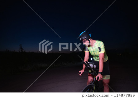 A triathlete resting on the road after a tough bike ride in the dark night, leaning on his bike in complete exhaustion A triathlete resting on the road after a tough bike ride in the dark night, leaning on his bike in complete exhaustion 114977989
