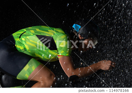A triathlete braving the rain as he cycles through the night, preparing himself for the upcoming marathon. The blurred raindrops in the foreground and the dark, moody atmosphere in the background add 114978166