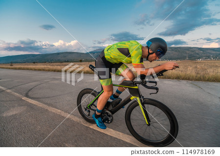 Triathlete riding his bicycle during sunset, preparing for a marathon. The warm colors of the sky provide a beautiful backdrop for his determined and focused effort. Triathlete riding his bicycle during sunset, preparing for a marathon. The warm colors of the sky provide a beautiful backdrop for his determined and focused effort. 114978169