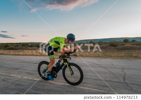Triathlete riding his bicycle during sunset, preparing for a marathon. The warm colors of the sky provide a beautiful backdrop for his determined and focused effort. 114978213