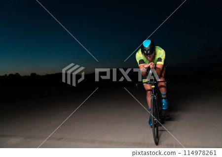 A triathlete rides his bike in the darkness of night, pushing himself to prepare for a marathon. The contrast between the darkness and the light of his bike creates a sense of drama and highlights the 114978261