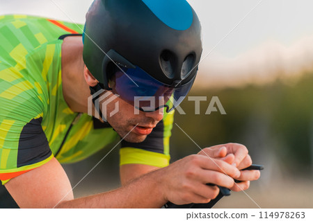 Close up photo of triathlete riding his bicycle during sunset, preparing for a marathon. The warm colors of the sky provide a beautiful backdrop for his determined and focused effort. Close up photo of triathlete riding his bicycle during sunset, preparing for a marathon. The warm colors of the sky provide a beautiful backdrop for his determined and focused effort. 114978263