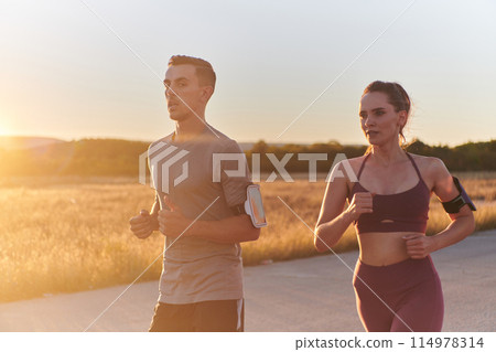 A handsome young couple running together during the early morning hours, with the mesmerizing sunrise casting a warm glow, symbolizing their shared love and vitality A handsome young couple running together during the early morning hours, with the mesmerizing sunrise casting a warm glow, symbolizing their shared love and vitality 114978314