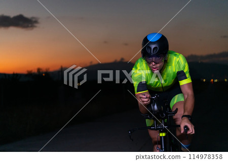 A triathlete rides his bike in the darkness of night, pushing himself to prepare for a marathon. The contrast between the darkness and the light of his bike creates a sense of drama and highlights the A triathlete rides his bike in the darkness of night, pushing himself to prepare for a marathon. The contrast between the darkness and the light of his bike creates a sense of drama and highlights the 114978358