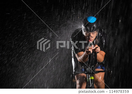 A triathlete braving the rain as he cycles through the night, preparing himself for the upcoming marathon. The blurred raindrops in the foreground and the dark, moody atmosphere in the background add A triathlete braving the rain as he cycles through the night, preparing himself for the upcoming marathon. The blurred raindrops in the foreground and the dark, moody atmosphere in the background add 114978359