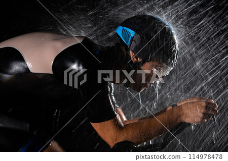 A triathlete braving the rain as he cycles through the night, preparing himself for the upcoming marathon. The blurred raindrops in the foreground and the dark, moody atmosphere in the background add 114978478