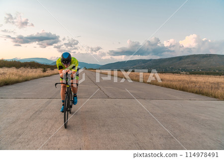 Triathlete riding his bicycle during sunset, preparing for a marathon. The warm colors of the sky provide a beautiful backdrop for his determined and focused effort. Triathlete riding his bicycle during sunset, preparing for a marathon. The warm colors of the sky provide a beautiful backdrop for his determined and focused effort. 114978491