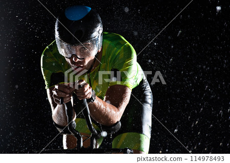 A triathlete braving the rain as he cycles through the night, preparing himself for the upcoming marathon. The blurred raindrops in the foreground and the dark, moody atmosphere in the background add 114978493