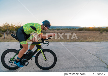 Triathlete riding his bicycle during sunset, preparing for a marathon. The warm colors of the sky provide a beautiful backdrop for his determined and focused effort. 114978547