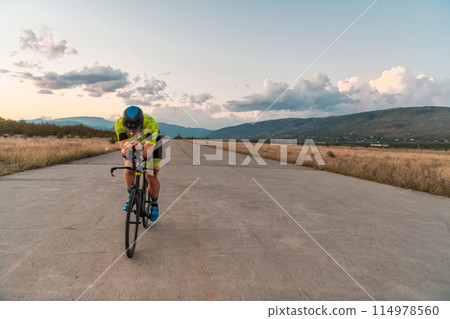 Triathlete riding his bicycle during sunset, preparing for a marathon. The warm colors of the sky provide a beautiful backdrop for his determined and focused effort. 114978560