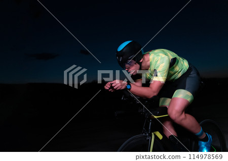A triathlete rides his bike in the darkness of night, pushing himself to prepare for a marathon. The contrast between the darkness and the light of his bike creates a sense of drama and highlights the A triathlete rides his bike in the darkness of night, pushing himself to prepare for a marathon. The contrast between the darkness and the light of his bike creates a sense of drama and highlights the 114978569