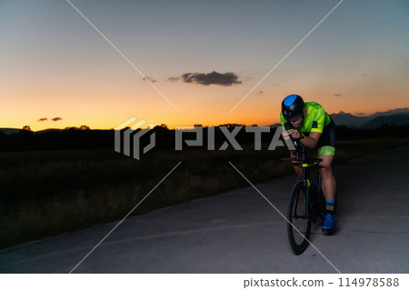 A triathlete rides his bike in the darkness of night, pushing himself to prepare for a marathon. The contrast between the darkness and the light of his bike creates a sense of drama and highlights the 114978588