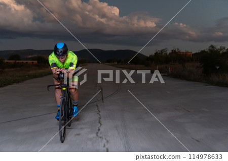 A triathlete rides his bike in the darkness of night, pushing himself to prepare for a marathon. The contrast between the darkness and the light of his bike creates a sense of drama and highlights the 114978633