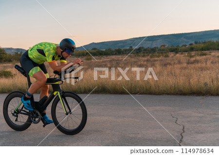 Triathlete riding his bicycle during sunset, preparing for a marathon. The warm colors of the sky provide a beautiful backdrop for his determined and focused effort. Triathlete riding his bicycle during sunset, preparing for a marathon. The warm colors of the sky provide a beautiful backdrop for his determined and focused effort. 114978634