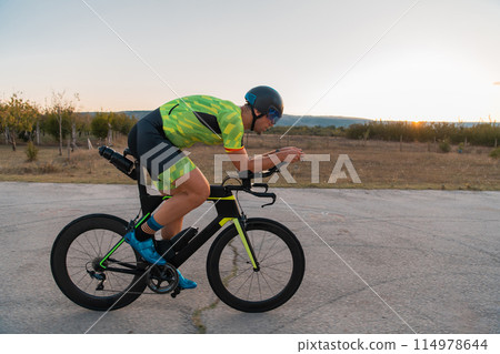 Triathlete riding his bicycle during sunset, preparing for a marathon. The warm colors of the sky provide a beautiful backdrop for his determined and focused effort. Triathlete riding his bicycle during sunset, preparing for a marathon. The warm colors of the sky provide a beautiful backdrop for his determined and focused effort. 114978644