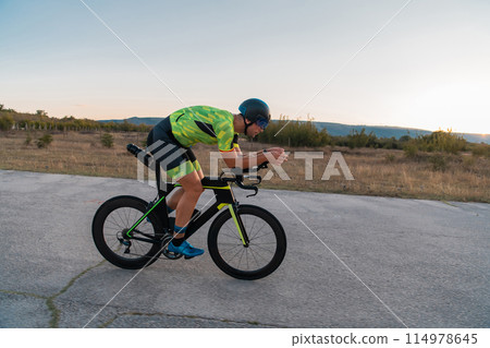 Triathlete riding his bicycle during sunset, preparing for a marathon. The warm colors of the sky provide a beautiful backdrop for his determined and focused effort. 114978645