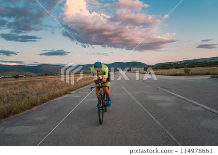 Triathlete riding his bicycle during sunset, preparing for a marathon. The warm colors of the sky provide a beautiful backdrop for his determined and focused effort. 114978661