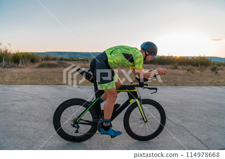 Triathlete riding his bicycle during sunset, preparing for a marathon. The warm colors of the sky provide a beautiful backdrop for his determined and focused effort. 114978668