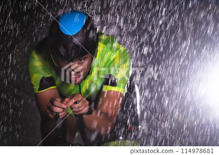 A triathlete braving the rain as he cycles through the night, preparing himself for the upcoming marathon. The blurred raindrops in the foreground and the dark, moody atmosphere in the background add 114978681