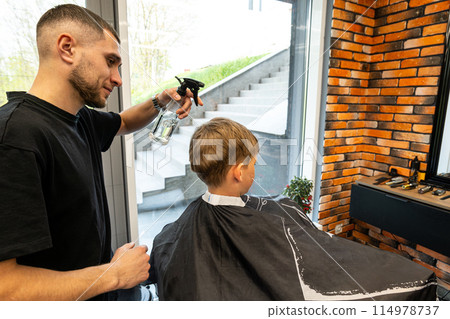 A boy sits in a barbershop while he is sprayed with water to start a haircut. A master in a barbershop sprays water on a boy who is sitting and waiting for his haircut. A boy sits in a barbershop while he is sprayed with water to start a haircut. A master in a barbershop sprays water on a boy who is sitting and waiting for his haircut. 114978737