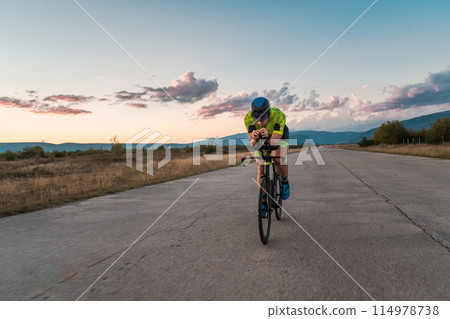 Triathlete riding his bicycle during sunset, preparing for a marathon. The warm colors of the sky provide a beautiful backdrop for his determined and focused effort. Triathlete riding his bicycle during sunset, preparing for a marathon. The warm colors of the sky provide a beautiful backdrop for his determined and focused effort. 114978738