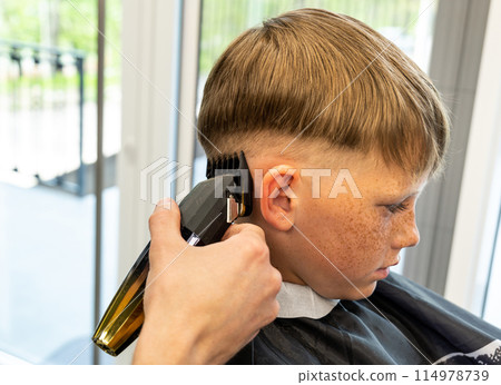 Coiffeur shaves client hair with electrical trimmer in modern barbershop closeup. Barber does elegant haircut contour in beauty salon Coiffeur shaves client hair with electrical trimmer in modern barbershop closeup. Barber does elegant haircut contour in beauty salon 114978739